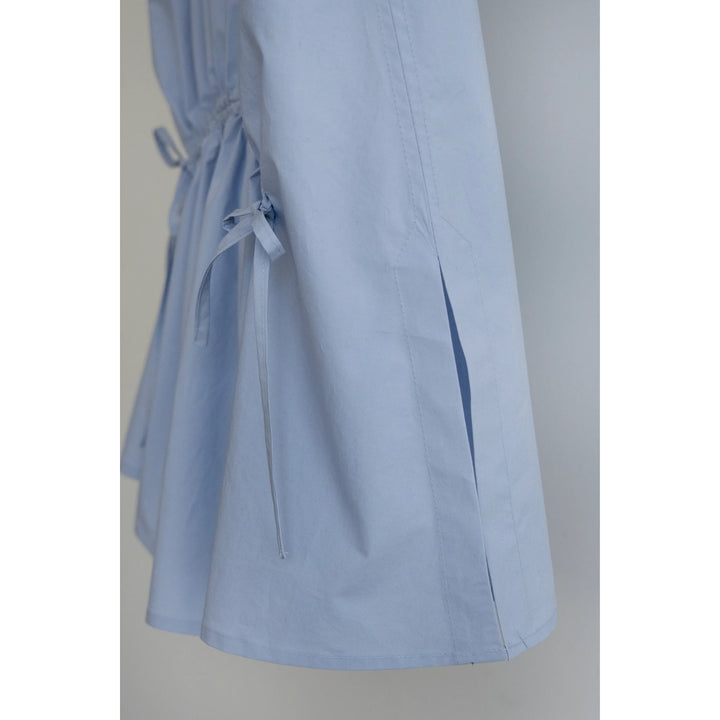 A close-up of a light blue garment, likely a dress or coat, featuring a drawstring waist and side slit details, shown against a neutral gray background.