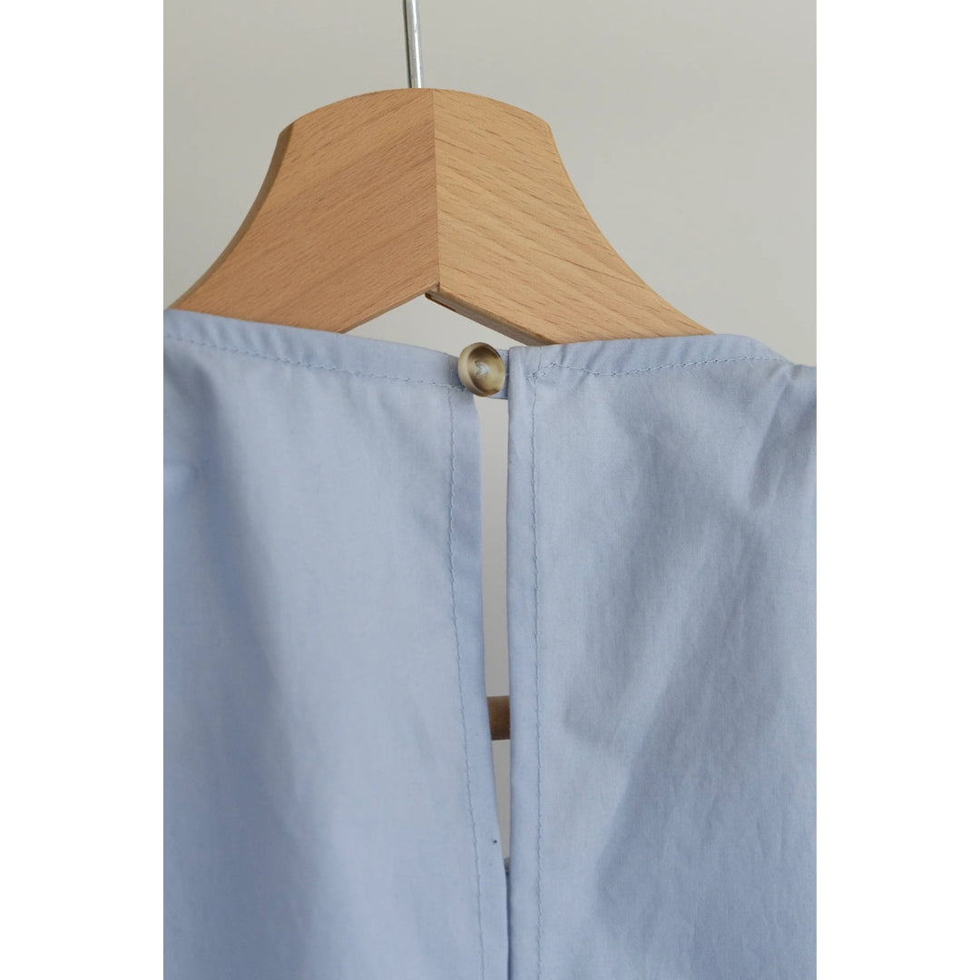 Close-up of a light blue garment hanging on a wooden hanger, showing the back neckline with a single gold-tone button and stitched panel seams.