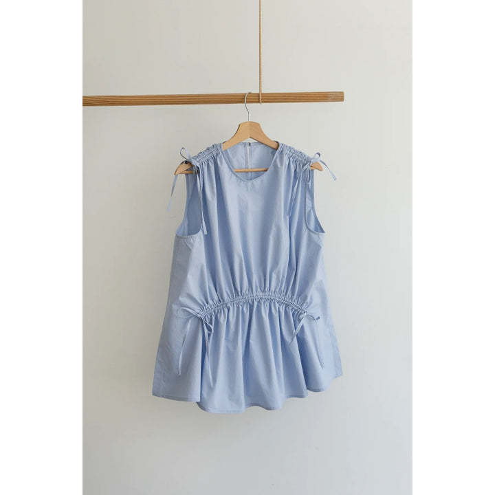 A light blue sleeveless top with shoulder tie details and a gathered waist, hanging on a wooden hanger from a wooden rod against a plain off-white wall.
