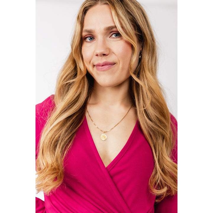A woman with long wavy blonde hair wearing a bright pink wrap top and a delicate gold necklace with a round pendant, photographed against a light gray background.