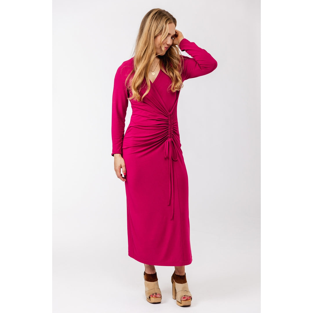 A woman modeling a long-sleeve magenta midi dress with a ruched wrap-style bodice and drawstring tie, paired with tan platform sandals, against a plain light gray studio background.