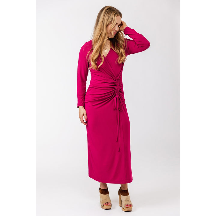 A woman modeling a long-sleeve magenta midi dress with a ruched wrap-style bodice and drawstring tie, paired with tan platform sandals, against a plain light gray studio background.