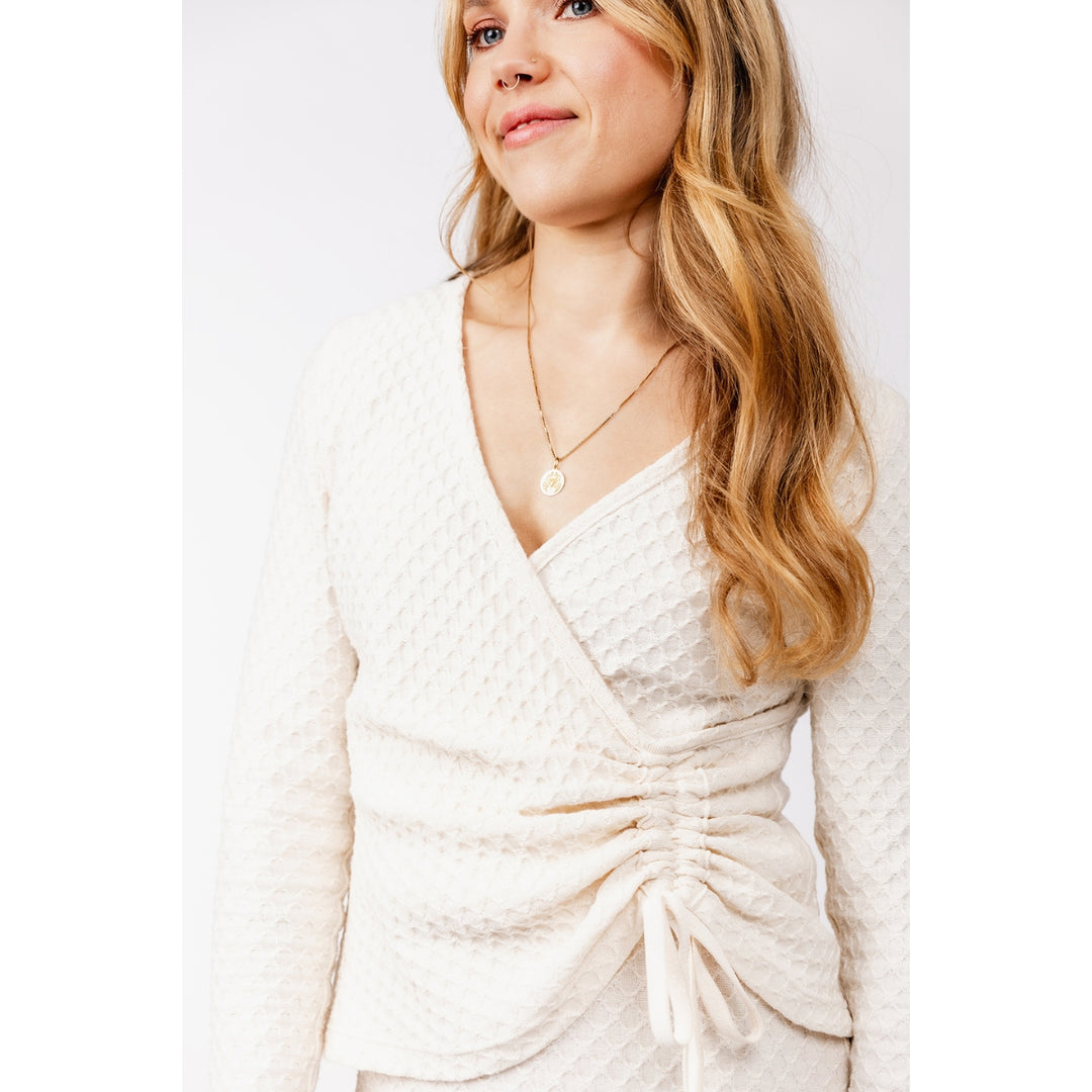 A woman wearing a cream-colored textured knit wrap top with a ruched waist and tie detail, shown against a plain light background.