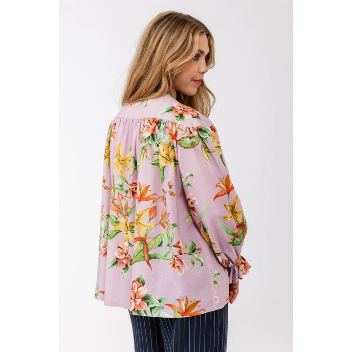 A woman shown from the back wearing a light pink long-sleeve blouse with a vibrant tropical floral print in orange, yellow, and green, paired with navy pinstriped trousers, against a plain light gray background.