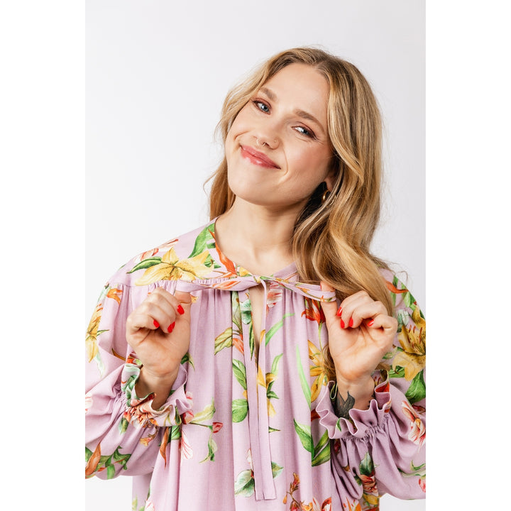 A woman with blonde hair wearing a light purple floral blouse with long ruffled sleeves and a tie-neck detail, posing against a light gray background.