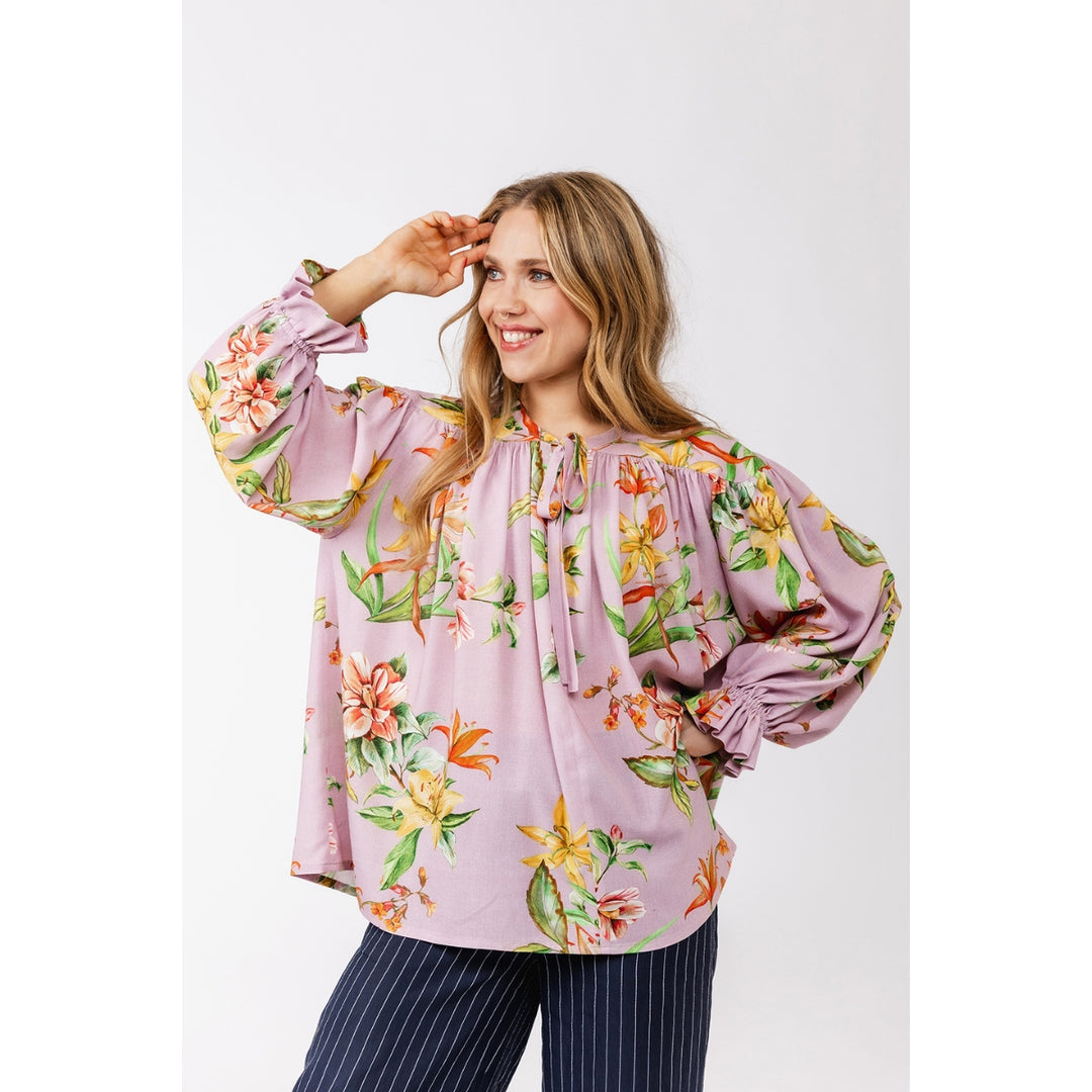 A woman modeling a lavender long-sleeve blouse with a vibrant orange, yellow, and green tropical floral print, paired with navy blue pinstriped trousers, against a plain light gray background.