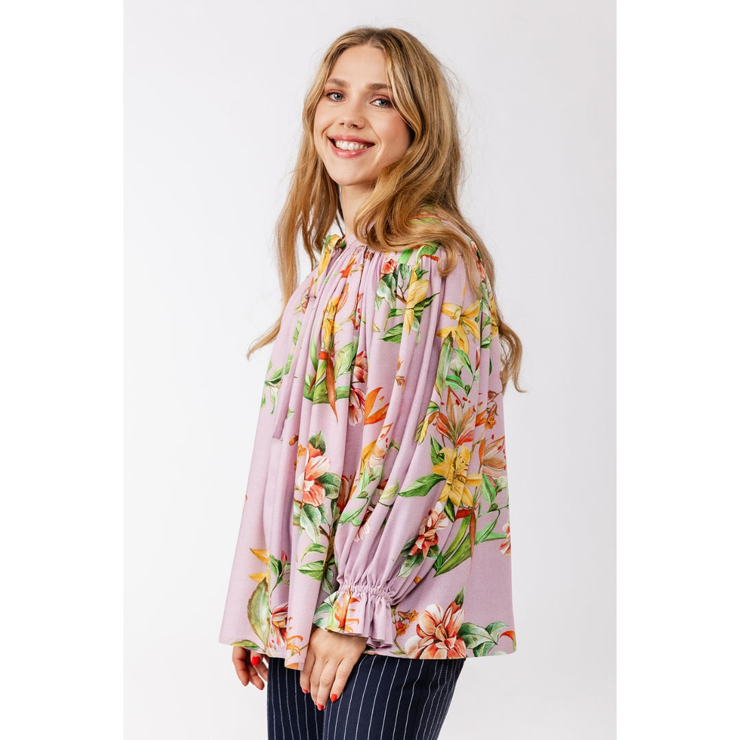 A woman modeling a light pink long-sleeve blouse with a vibrant orange, yellow, and green tropical floral print, paired with navy blue pinstriped trousers, against a plain light gray background.
