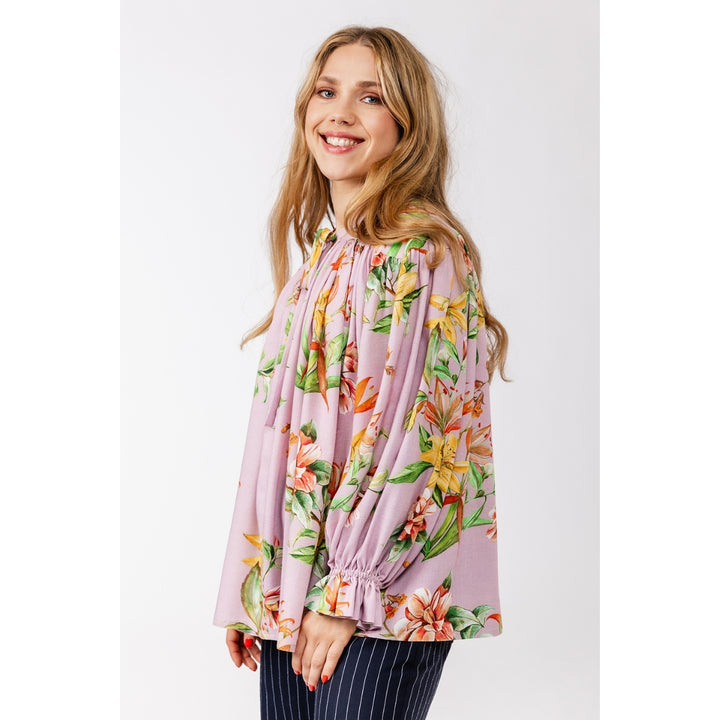 A woman modeling a light pink long-sleeve blouse with a vibrant orange, yellow, and green tropical floral print, paired with navy blue pinstriped trousers, against a plain light gray background.
