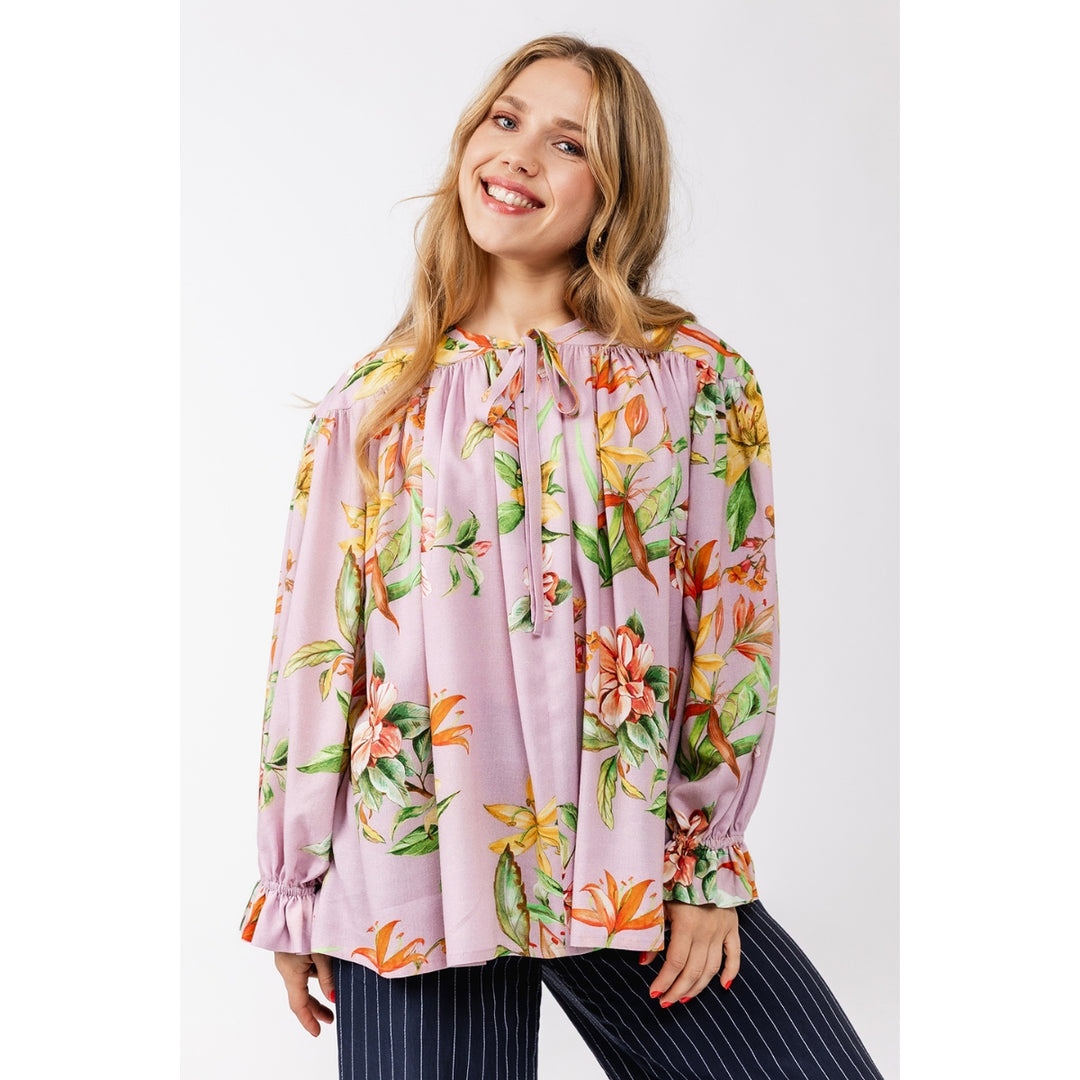 A woman wearing a light pink long-sleeve blouse with a vibrant tropical floral print in orange, yellow, and green, paired with navy blue pinstriped pants, set against a plain light gray studio background.