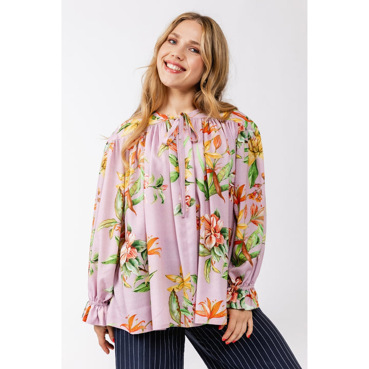 A woman wearing a light pink long-sleeve blouse with a vibrant tropical floral print in orange, yellow, and green, paired with navy blue pinstriped pants, set against a plain light gray studio background.