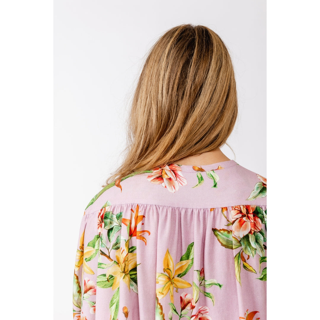 A woman shown from the back wearing a light pink floral blouse with yellow, orange, and green tropical flowers, set against a plain white background.
