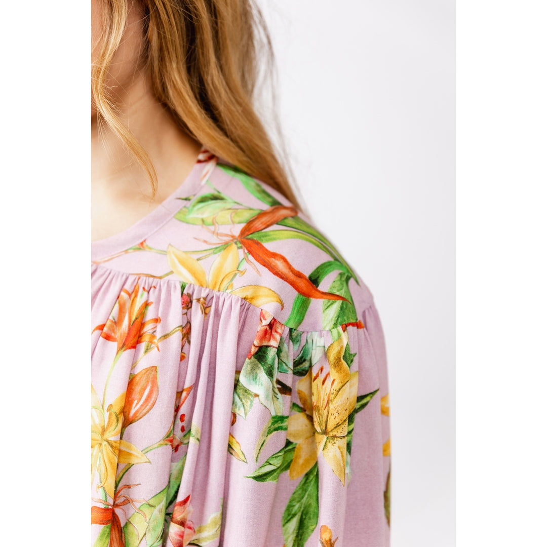 Close-up of a woman wearing a light pink floral top with yellow, orange, and green botanical print against a plain white background.