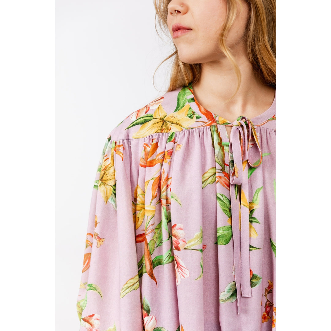A close-up of a model wearing a lavender floral blouse with a tie-neck detail, featuring yellow, orange, and green botanical patterns.