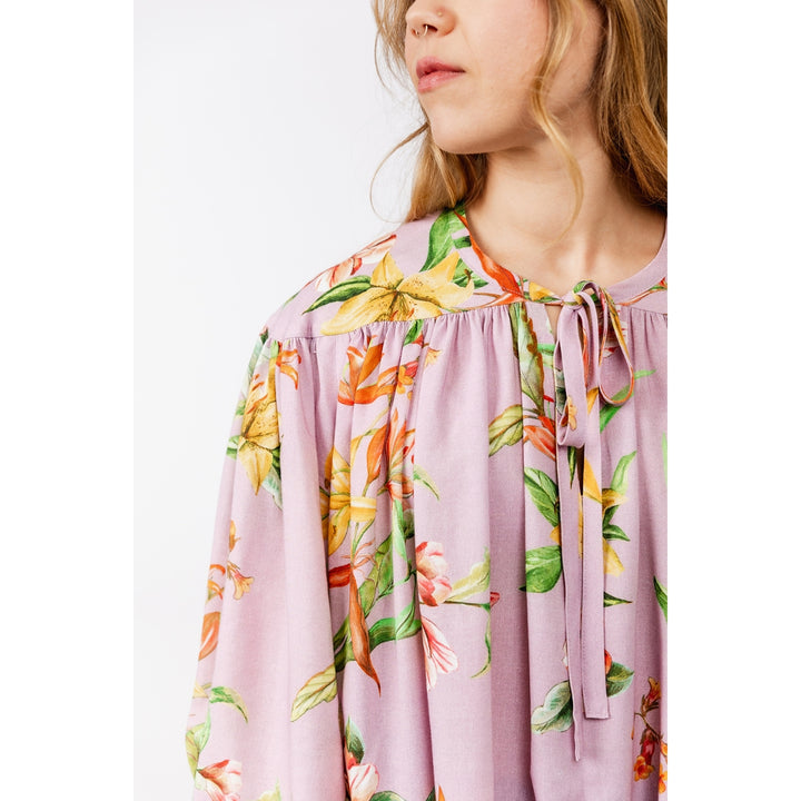 A close-up of a model wearing a lavender floral blouse with a tie-neck detail, featuring yellow, orange, and green botanical patterns.