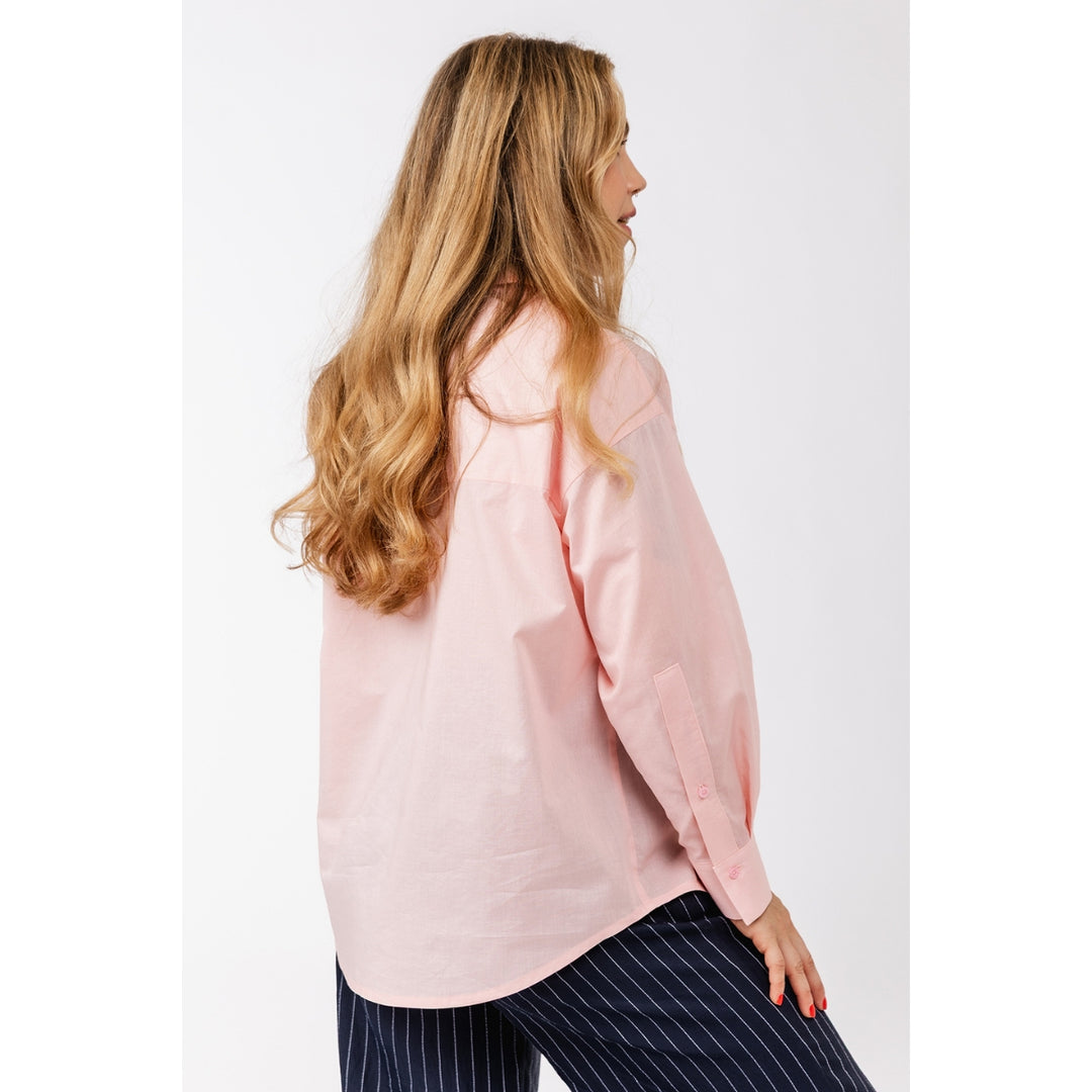 A woman with long blonde hair is shown from the back and side, wearing an oversized light pink button-up shirt and navy pinstriped pants against a plain light gray studio background.