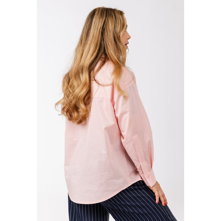 A woman with long blonde hair is shown from the back and side, wearing an oversized light pink button-up shirt and navy pinstriped pants against a plain light gray studio background.