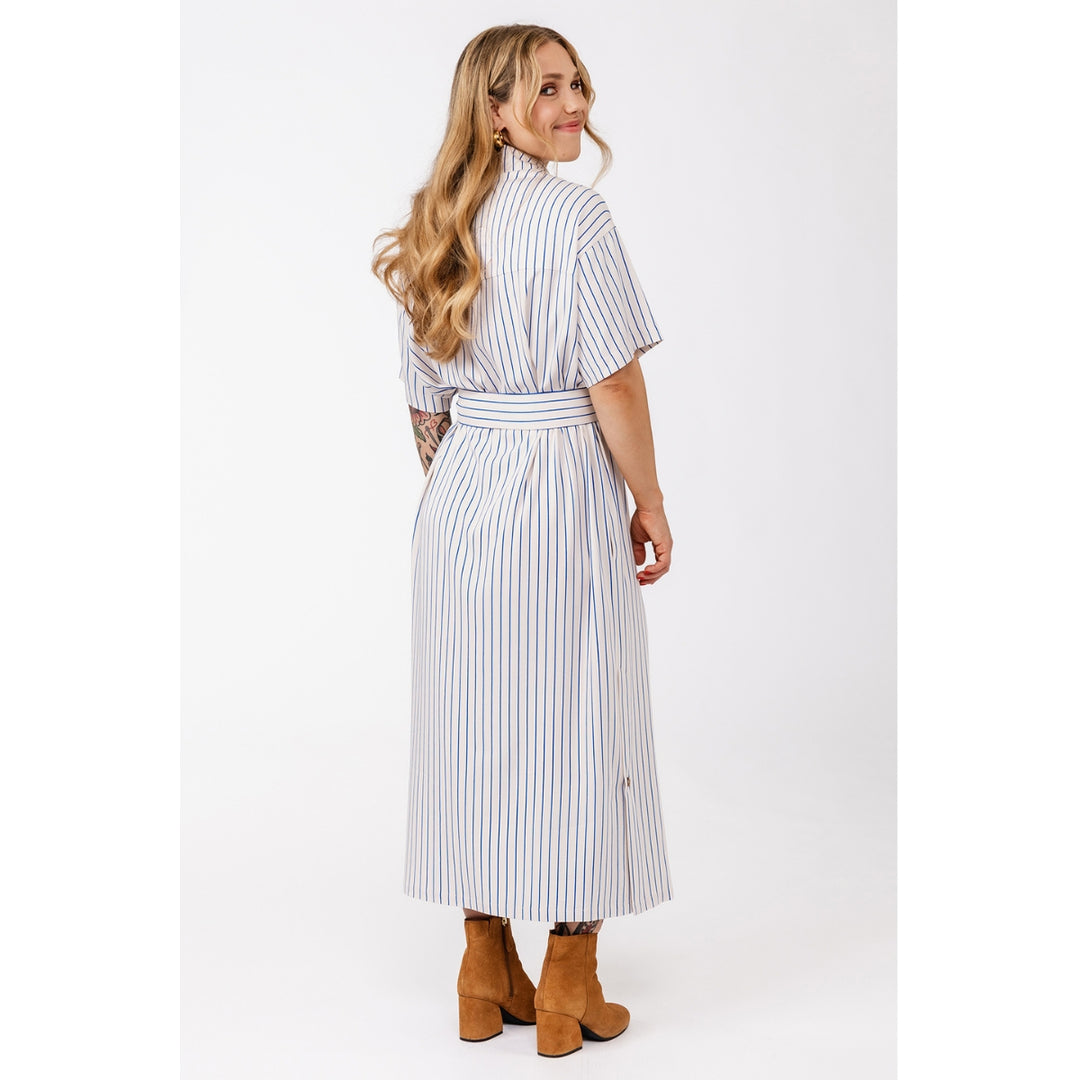 A woman modeling a white midi shirt dress with thin blue vertical stripes, featuring a high collar, short sleeves, and a matching fabric tie belt. She is shown from the back, wearing brown ankle boots, against a plain white studio background.