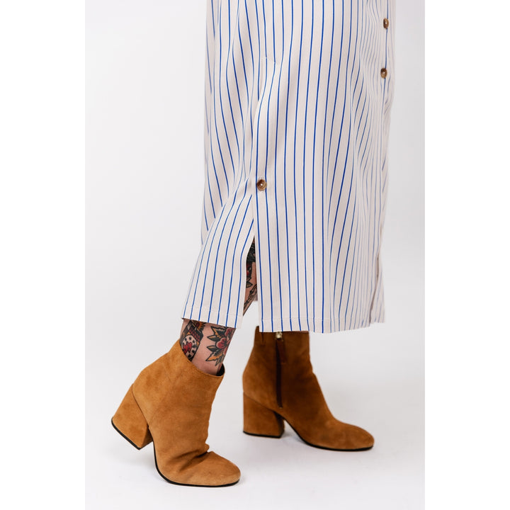 A person wearing brown suede block-heel ankle boots with a white and blue vertically striped shirt skirt, shown from the waist down against a light gray studio background.