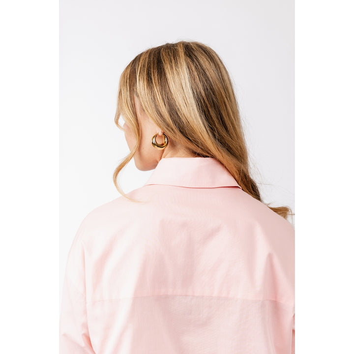 A woman with blonde-highlighted hair is shown from the back wearing a light pink button-up shirt and a gold hoop earring, photographed against a plain white studio background.