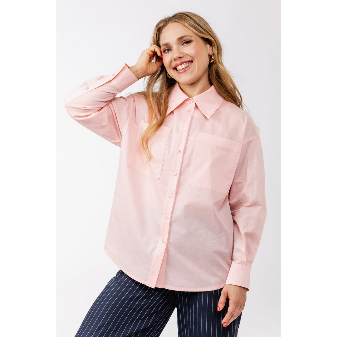 A woman modeling a light pink long-sleeve button-up shirt with a pointed collar and chest pocket, paired with navy pinstripe pants, against a plain light gray studio background.