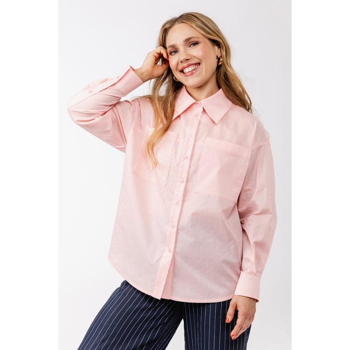 A woman modeling a light pink long-sleeve button-up shirt with a pointed collar and chest pocket, paired with navy pinstripe pants, against a plain light gray studio background.