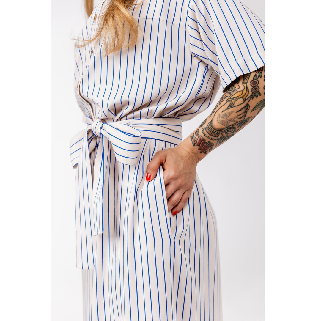 A close-up of a person wearing a white belted jumpsuit with thin blue vertical stripes. The person has a tattooed arm and red nail polish, and the background is a clean white studio backdrop.