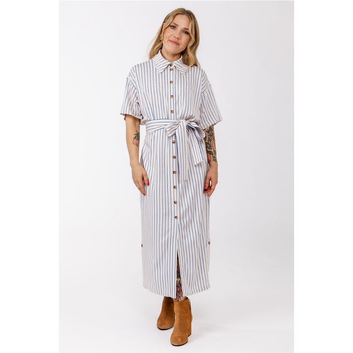 A woman modeling a white short-sleeve midi shirt dress with thin blue vertical stripes, a matching tie waist belt, and brown suede ankle boots against a light gray studio background.