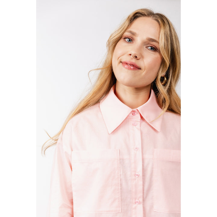A woman with blonde hair wearing a light pink button-up shirt with a pointed collar and two chest pockets, photographed against a plain light gray studio background.