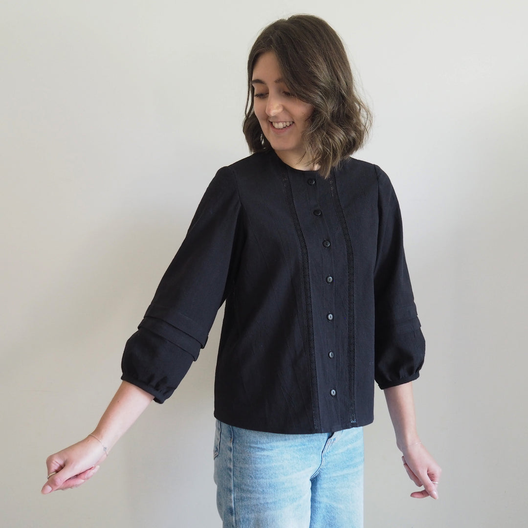 Collarless Nepheline blouse in black textured cotton with delicate lace panel down the front of the button up blouse and pleated sleeves.