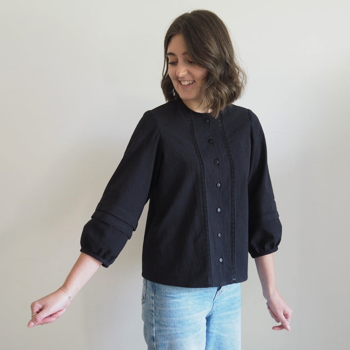 Collarless Nepheline blouse in black textured cotton with delicate lace panel down the front of the button up blouse and pleated sleeves.