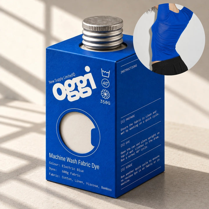 A blue Oggi brand machine wash fabric dye carton with a silver metal cap, shown on a light surface with soft window shadows. A circular inset shows a person wearing a bright blue top, demonstrating the dye result.