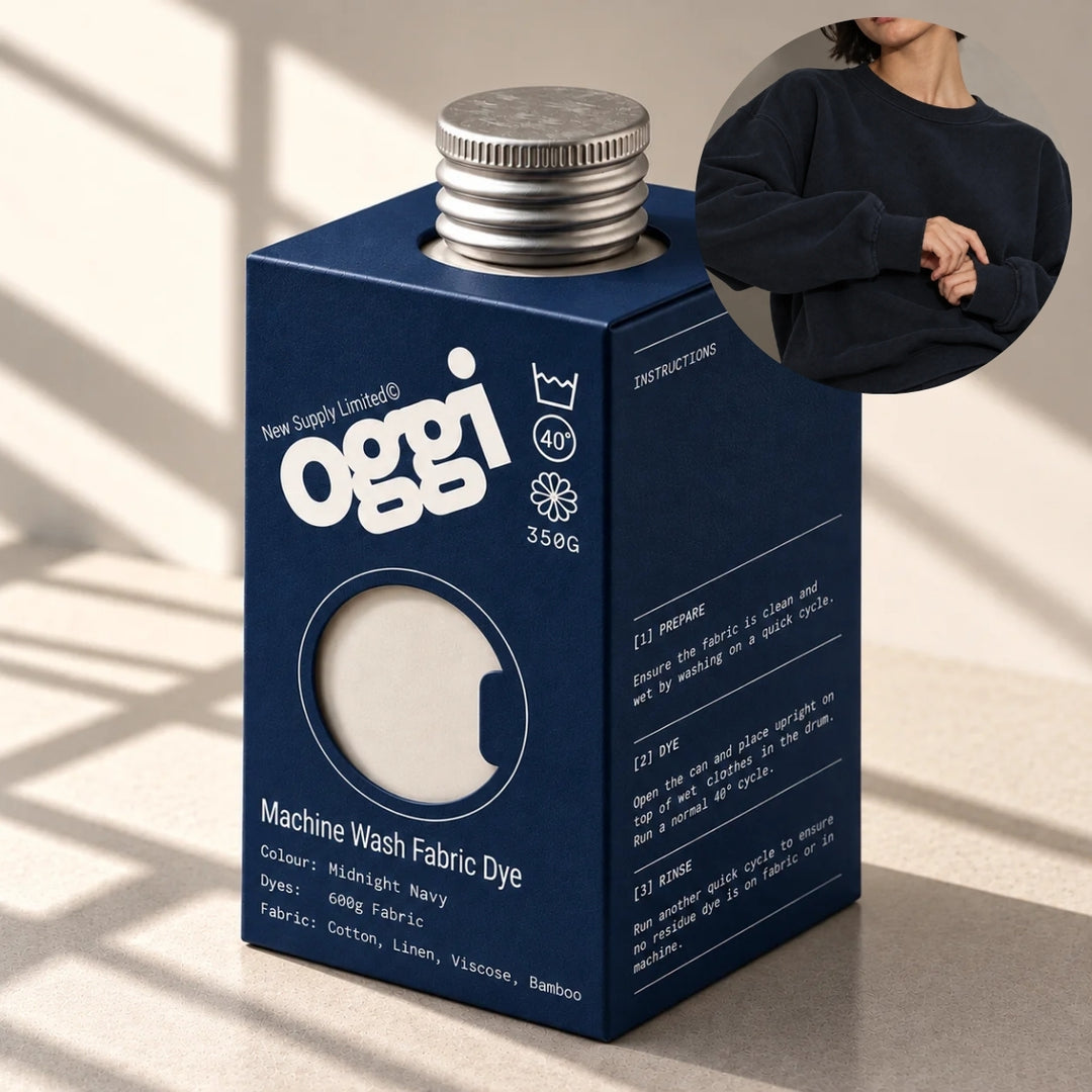 A dark blue Oggi brand box of Machine Wash Fabric Dye, shown on a light beige surface with soft window shadows. A circular inset in the top right shows a person wearing a navy sweatshirt to demonstrate the dye result.