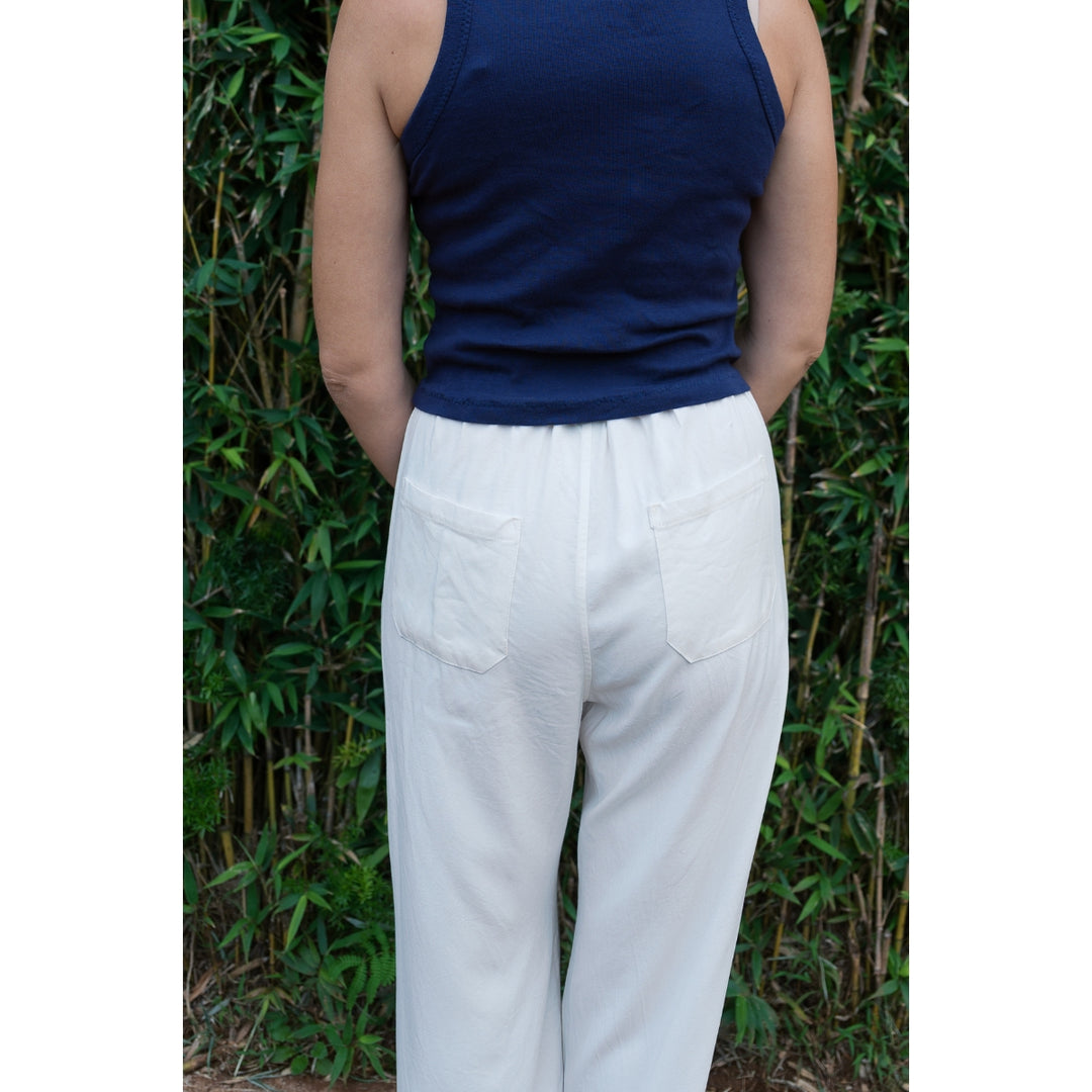 Person wearing a navy top and white pants standing against a green leafy background
