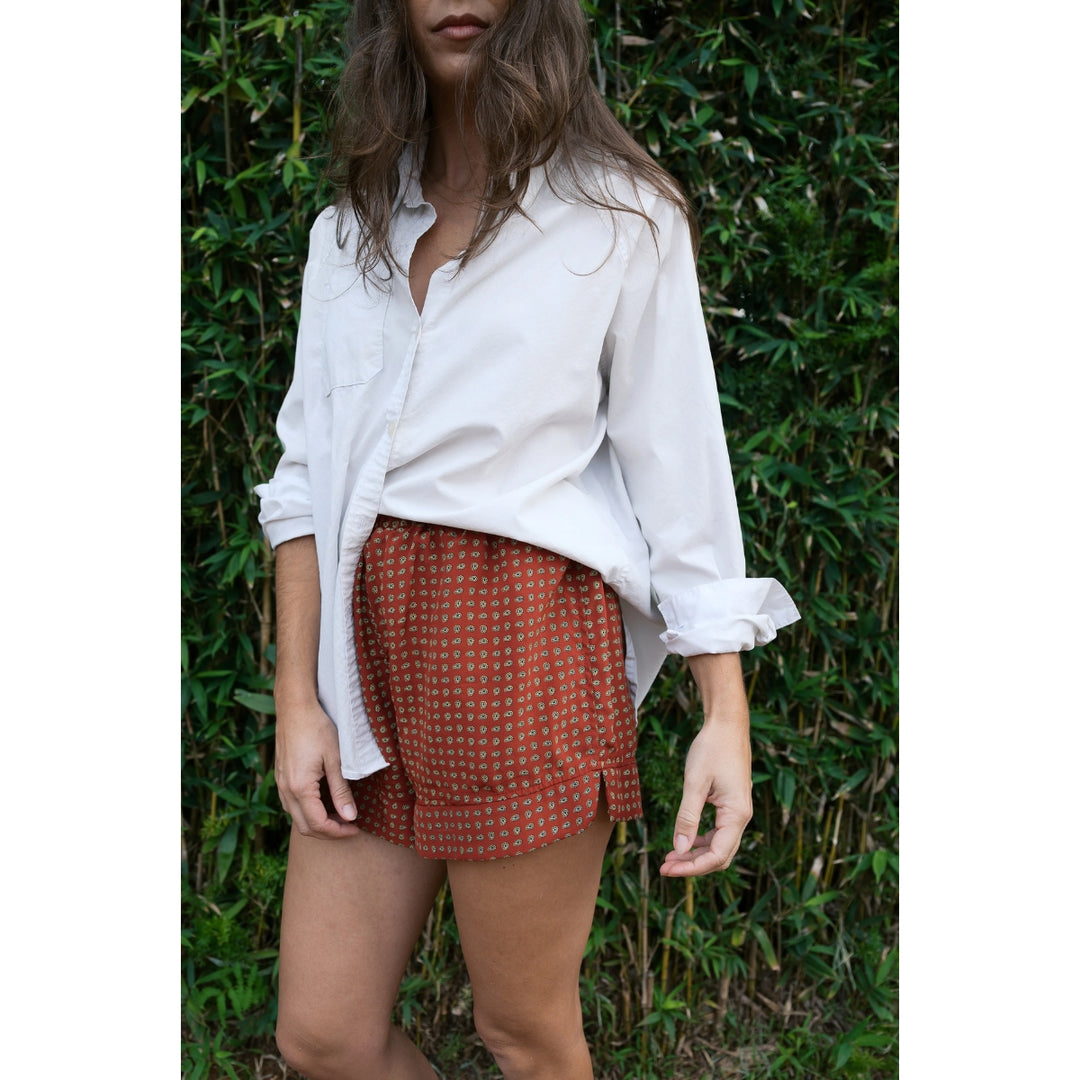 Person wearing a white shirt and patterned shorts standing against a green leafy background