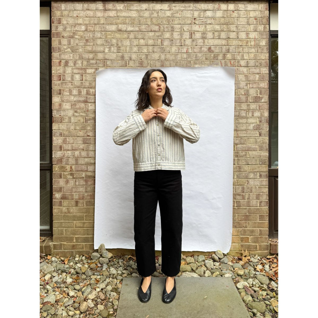 Person wearing a white striped shirt and black pants standing against a white backdrop in front of a brick wall.