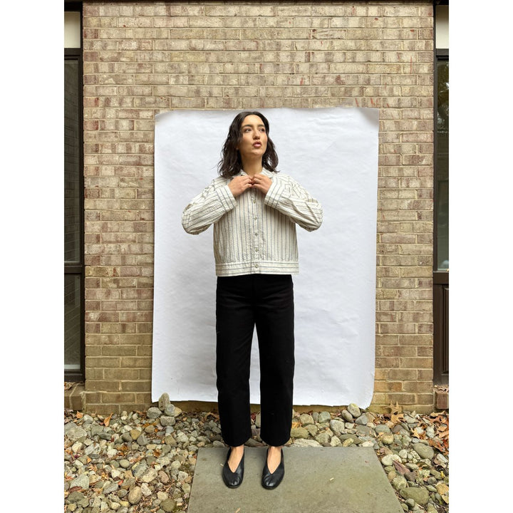 Person wearing a white striped shirt and black pants standing against a white backdrop in front of a brick wall.