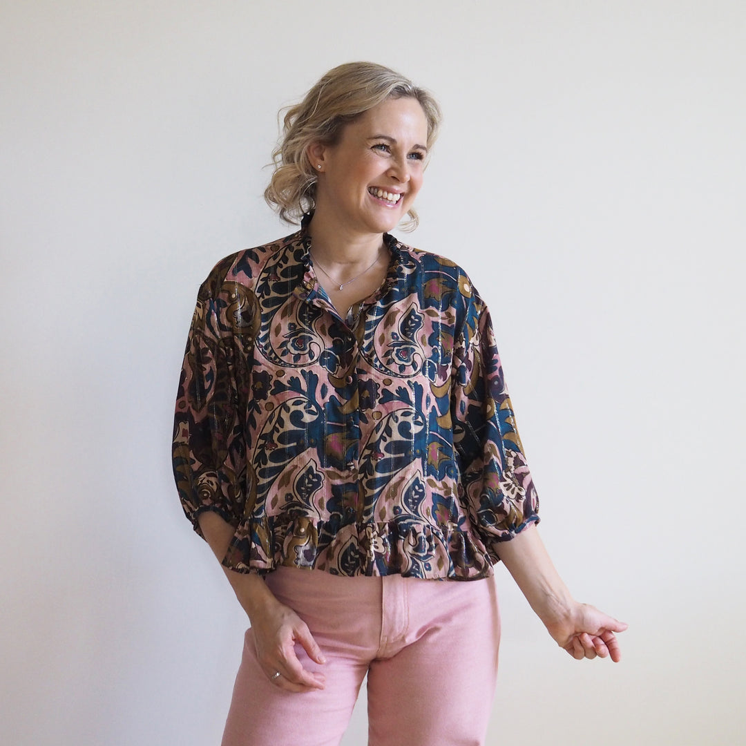 A woman wearing a patterned button-up blouse with three-quarter sleeves and pink pants, standing against a plain off-white wall.