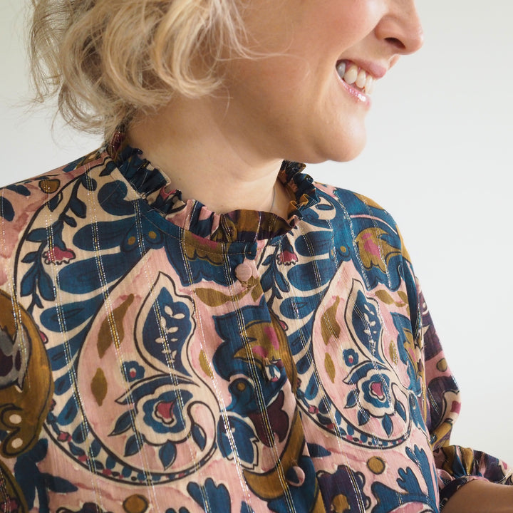 A close-up of a woman wearing a long-sleeve blouse with a ruffled high neck and a detailed paisley pattern in dusty pink, navy blue, and brown.