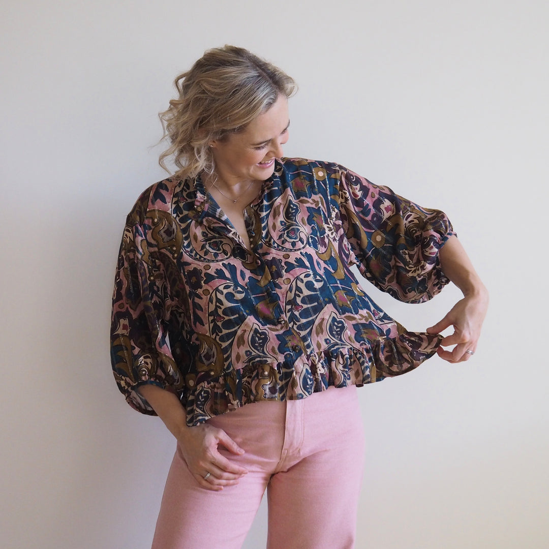 A woman wearing a loose, long-sleeve blouse with a dark brown, pink, and blue abstract pattern, paired with light pink pants, standing against a plain off-white wall.