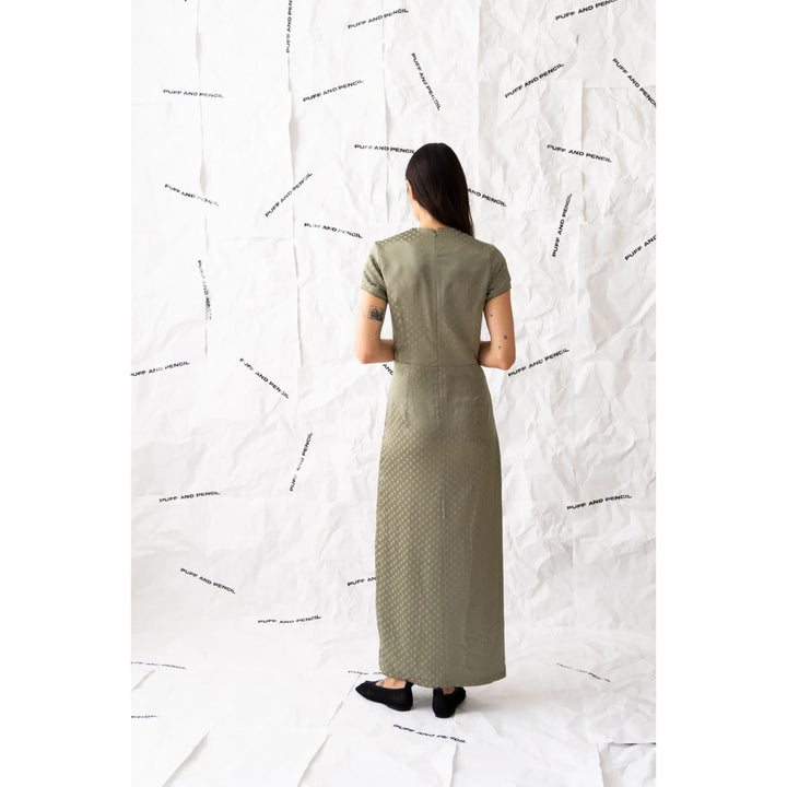 Woman wearing a long green dress standing against a white wall with black abstract patterns.