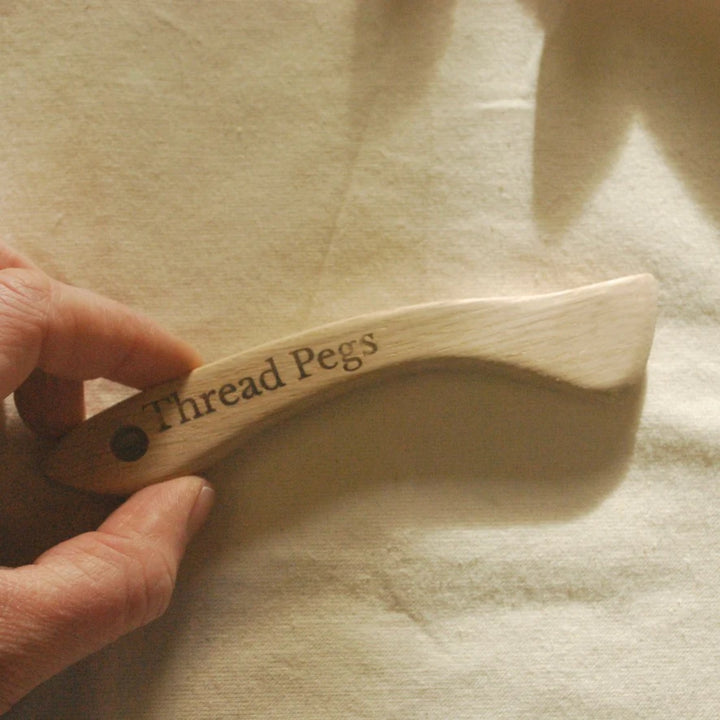 A hand holding a curved wooden sewing tool engraved with the words 'Thread Pegs' on a beige fabric background.