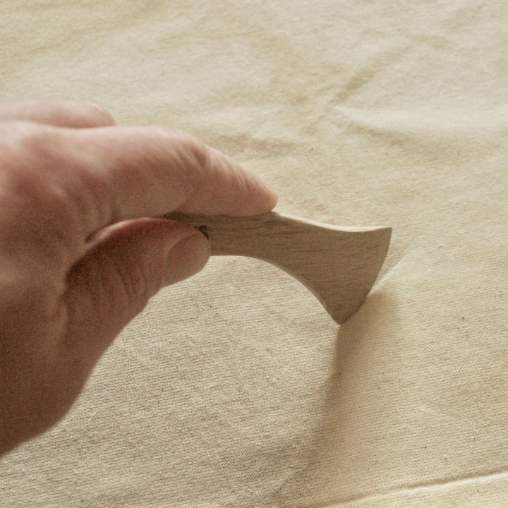 A hand holding a small wooden sewing tool with a light natural wood handle, shown against a neutral beige fabric background.