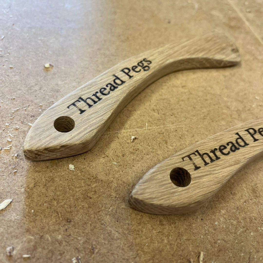 Two curved wooden thread pegs with pre-drilled holes, engraved with the text 'Thread Pegs', displayed on a brown wood-grain surface.