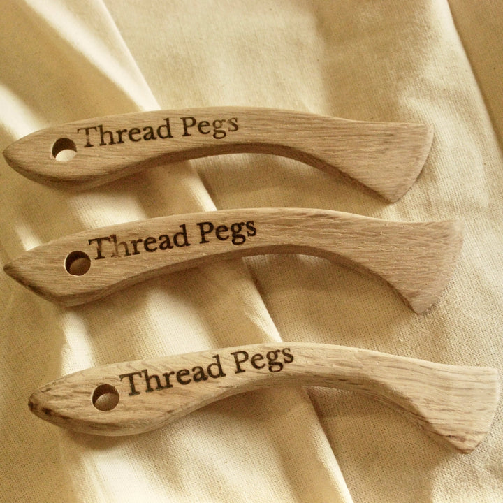 Three wooden thread pegs with 'Thread Pegs' engraving on a beige fabric background