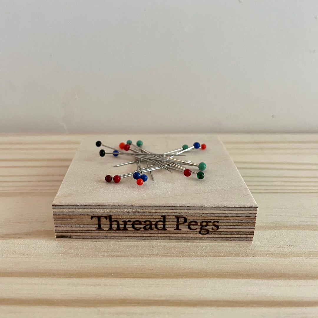 A small wooden tool engraved with the words 'Thread Pegs' sits on a light wood surface, with several pins featuring colourful tips resting on top.