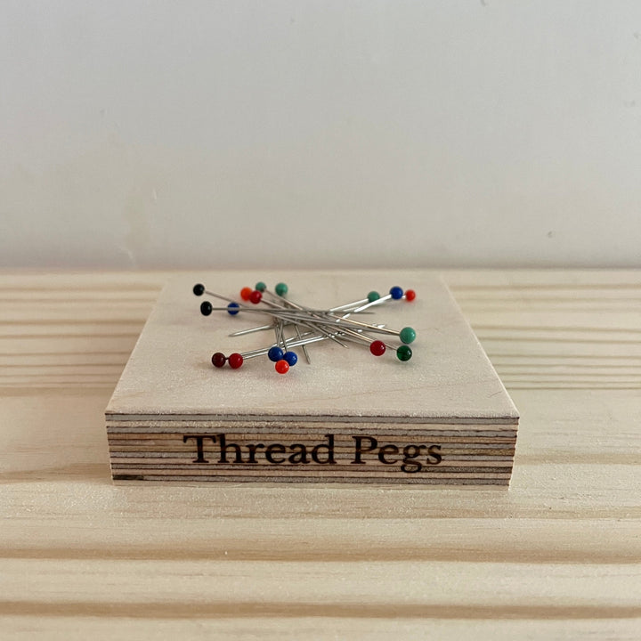 A small wooden tool engraved with the words 'Thread Pegs' sits on a light wood surface, with several pins featuring colourful tips resting on top.