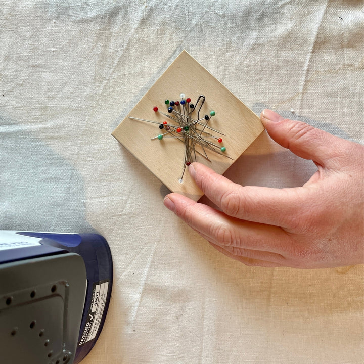 A hand holding a small square wooden pin magnetic covered with assorted sewing pins, set on a beige fabric surface with part of a blue iron visible in the lower left corner.