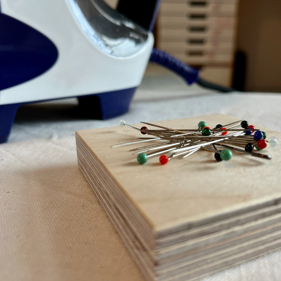 A close-up of a wooden pin magnet holding several metal sewing pins with colourful plastic tips, with a blurred iron in the background on a fabric surface.