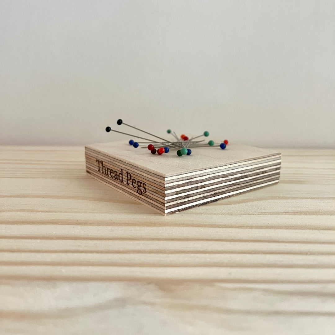 A small wooden box labeled 'Thread Pegs' holding several sewing pins, placed on a light wood tabletop against a plain light wall.