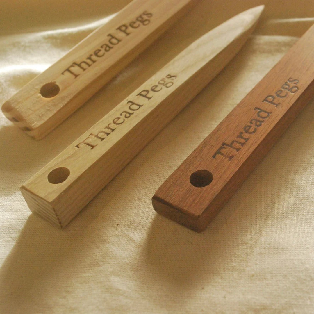 Three wooden thread pegs with engraved text 'Thread Pegs' and a hole at one end, shown on a beige fabric background. Two are light natural wood and one is medium brown wood.